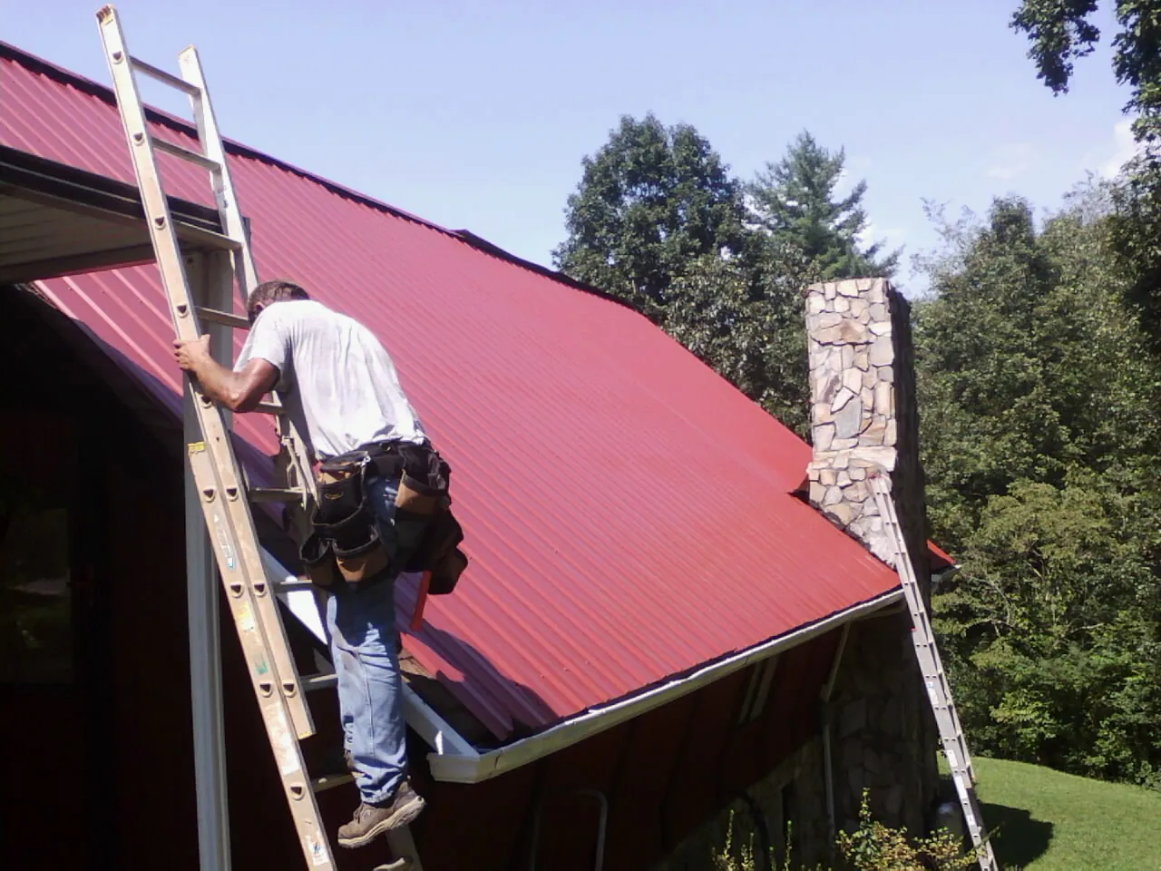Roofer on a ladder beside a red metal roof and stone chimney — Soffit & Fascia Repair by Roofing Brothers in Lykens
